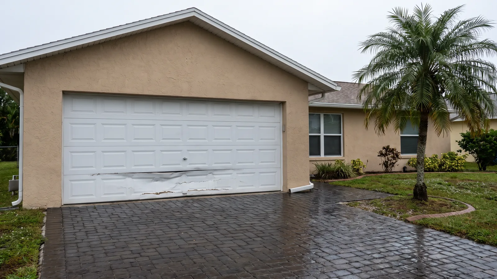 White garage door with its lowest section creased and dented where a car tapped it, on a tan ranch home in Port St. Lucie