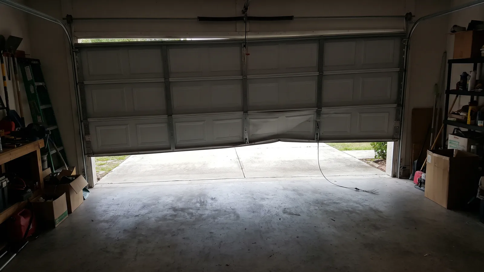 Sectional garage door hanging crooked in its opening with a frayed lift cable dangling, seen from inside a Florida garage toward a bright driveway